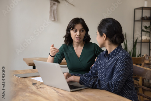 Two young office workers, female students sit at desk with laptop, share ideas, talk, discuss business project, brainstorm, analyze new joint task, use modern technology for work, engaged in teamwork