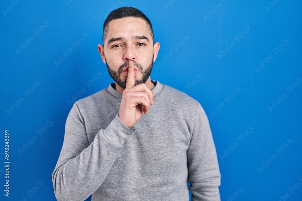 Hispanic man standing over blue background asking to be quiet with finger on lips. silence and secret concept. Hispanic man standing over blue background asking to be quiet with finger on lips. silence and secret concept.