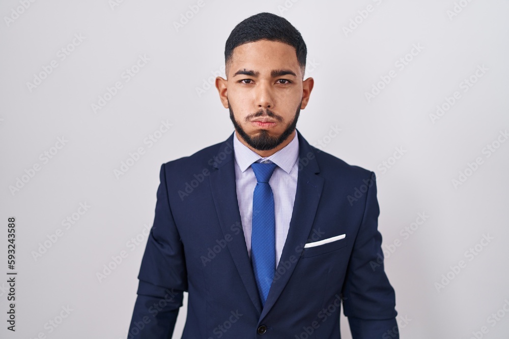 Young hispanic man wearing business suit and tie puffing cheeks with funny face. mouth inflated with air, crazy expression. Young hispanic man wearing business suit and tie puffing cheeks with funny face. mouth inflated with air, crazy expression.