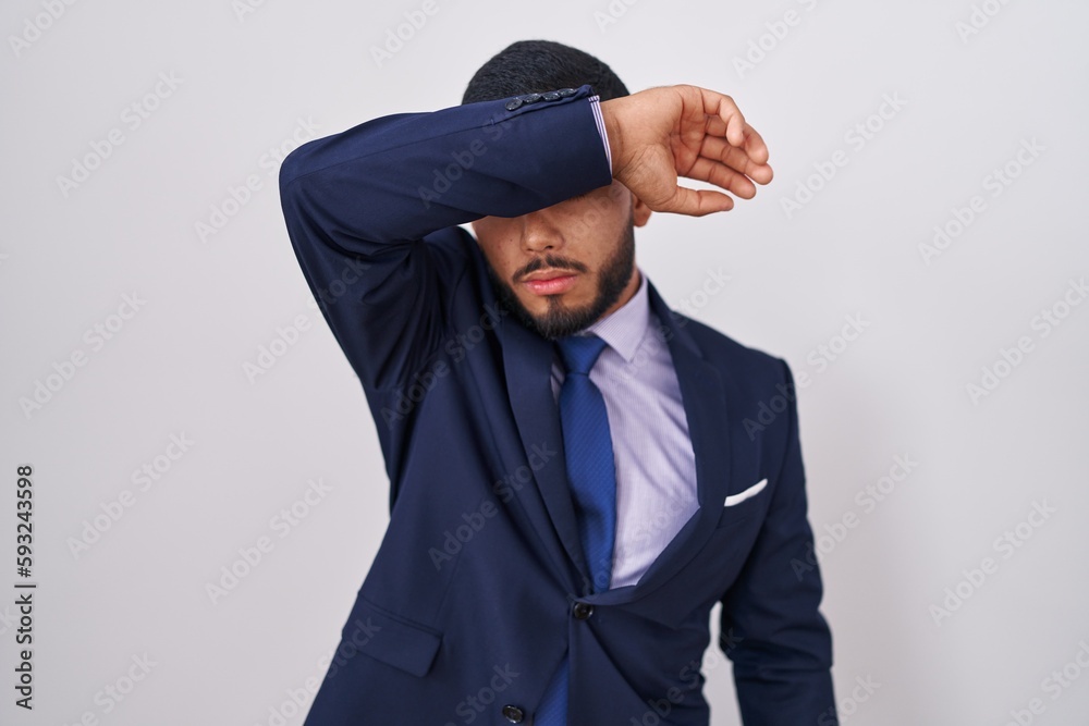 Young hispanic man wearing business suit and tie covering eyes with arm, looking serious and sad. sightless, hiding and rejection concept
