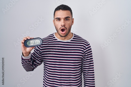 Young hispanic man wearing pyjama holding alarm clock scared and amazed with open mouth for surprise, disbelief face
