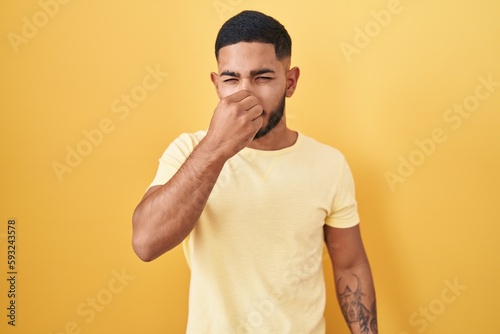 Young hispanic man standing over yellow background smelling something stinky and disgusting, intolerable smell, holding breath with fingers on nose. bad smell