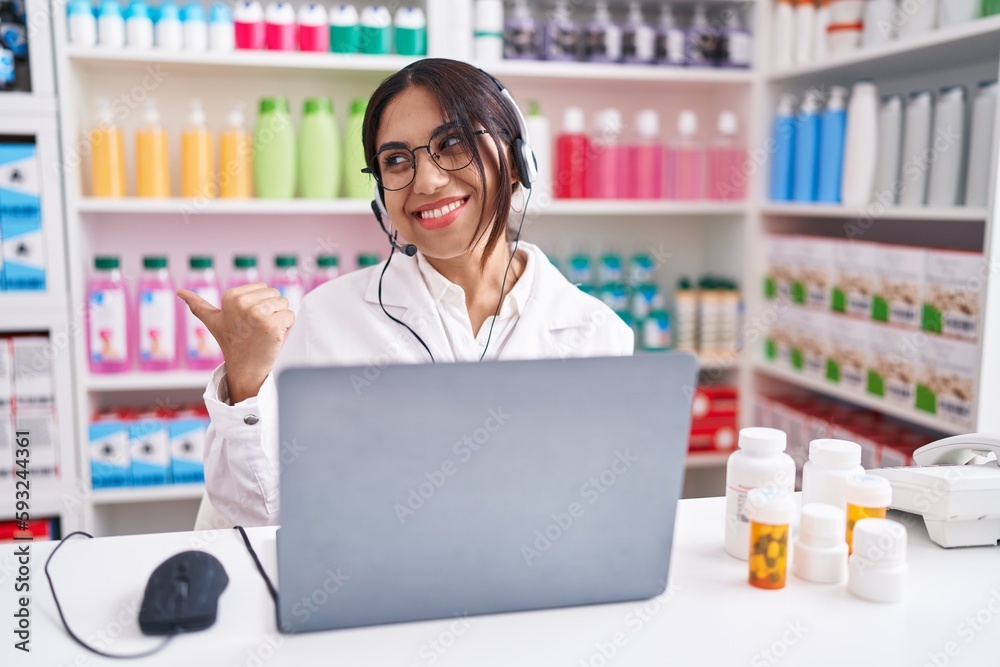 Young arab woman working at pharmacy drugstore using laptop smiling with happy face looking and pointing to the side with thumb up.