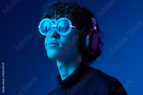 Teenage male with headphones listening to music and dancing and singing with glasses, hipster lifestyle, blue background, neon light, style and trends, mixed light, copy space