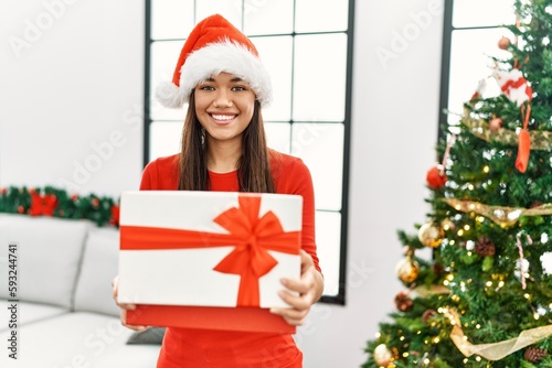 Young latin woman unboxing gift standing by christmas tree at home