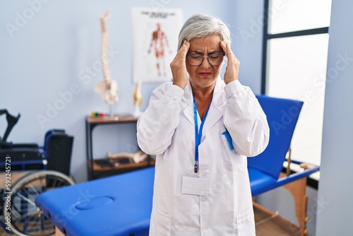 Middle age woman with grey hair working at pain recovery clinic with hand on head, headache because stress. suffering migraine.