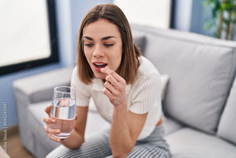 Young beautiful hispanic woman taking pill sitting on sofa at home Young beautiful hispanic woman taking pill sitting on sofa at home