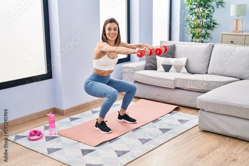 Young beautiful hispanic woman training leg exercise using dumbbells at home