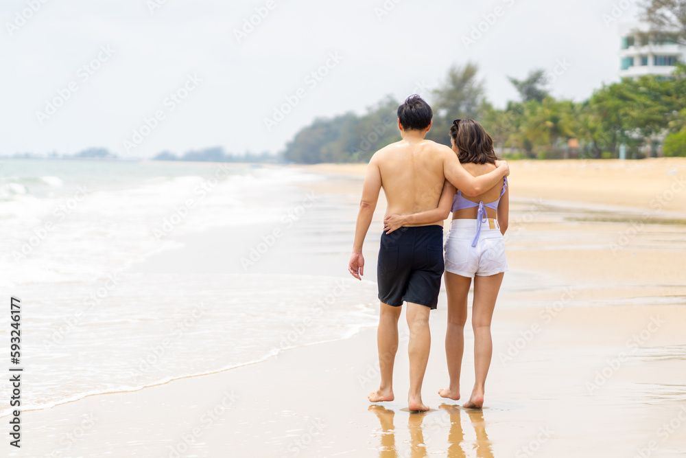 Happy Asian family couple in swimwear holding hands and walking together on tropical island beach at summer sunset. Husband and wife enjoy and fun outdoor lifestyle travel ocean on holiday vacation.