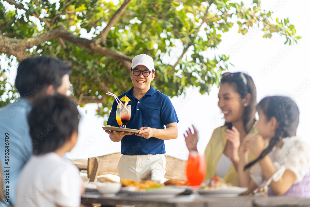 Happy Asian family couple and little kids travel ocean on summer holiday vacation. Waiter serving food and drink to customers on the table at beach restaurant at tropical island beach in sunny day. Happy Asian family couple and little kids travel ocean on summer holiday vacation. Waiter serving food and drink to customers on the table at beach restaurant at tropical island beach in sunny day.