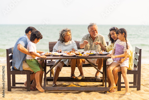 Happy Multi-Generation Asian family having celebration dinner food and drink together at tropical beach restaurant during travel ocean on summer holiday vacation at sunset. Family relationship concept