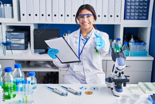 Hispanic young woman working at scientist laboratory smiling friendly offering handshake as greeting and welcoming. successful business.