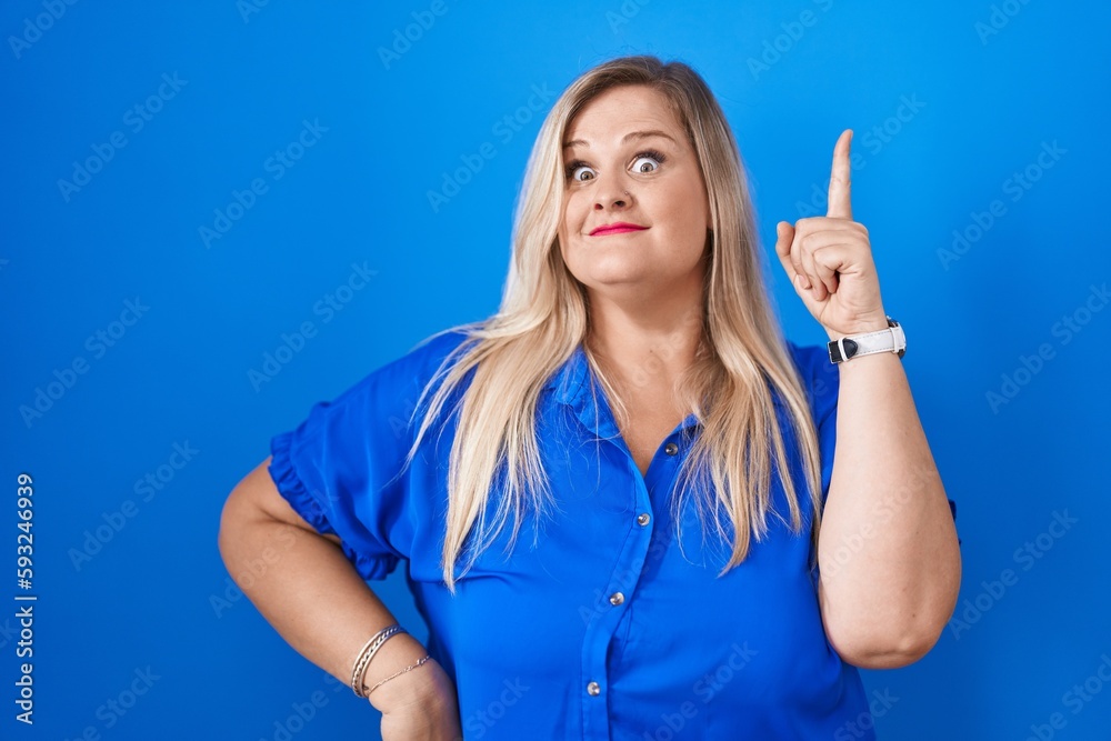 Caucasian plus size woman standing over blue background pointing finger up with successful idea. exited and happy. number one. Caucasian plus size woman standing over blue background pointing finger up with successful idea. exited and happy. number one.