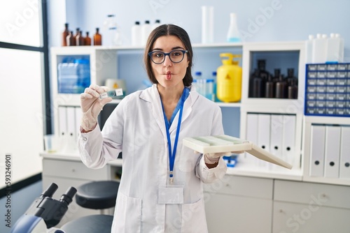 Young hispanic woman working at scientist laboratory with blood samples making fish face with mouth and squinting eyes  crazy and comical.
