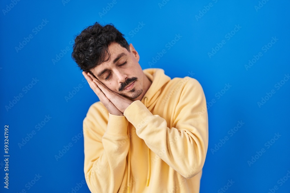 Hispanic man standing over blue background sleeping tired dreaming and posing with hands together while smiling with closed eyes. Hispanic man standing over blue background sleeping tired dreaming and posing with hands together while smiling with closed eyes.