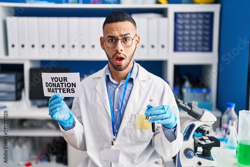 Young hispanic man working at scientist laboratory holding your donation matters holding blood sample clueless and confused expression. doubt concept.