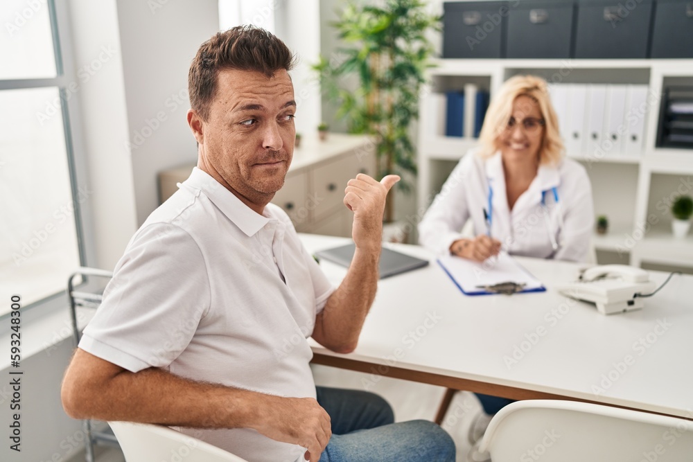 Hispanic man at the doctor pointing thumb up to the side smiling happy with open mouth