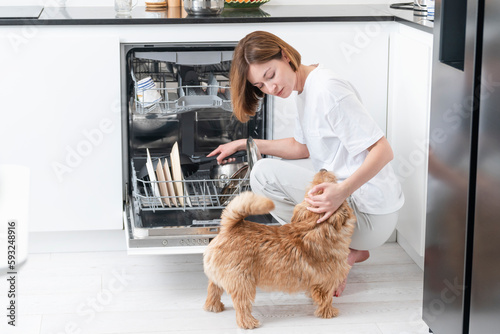 Woman loading dirty dishes inside dishwasher machine and her cute dog trying to play with her 