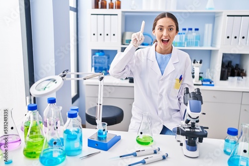 Young brunette woman working at scientist laboratory pointing finger up with successful idea. exited and happy. number one.