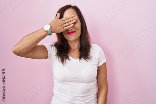 Middle age brunette woman standing over pink background smiling and laughing with hand on face covering eyes for surprise. blind concept.