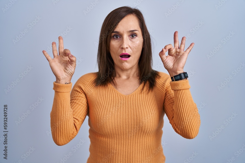Middle age brunette woman standing wearing orange sweater looking surprised and shocked doing ok approval symbol with fingers. crazy expression