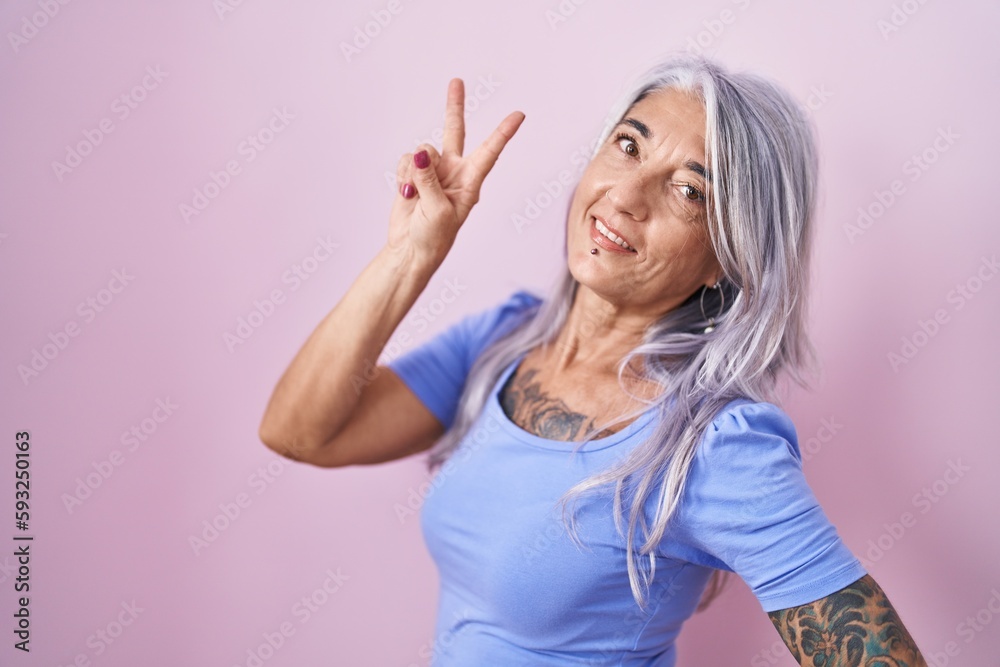 Fototapeta premium Middle age woman with tattoos standing over pink background smiling looking to the camera showing fingers doing victory sign. number two.