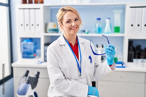 Young blonde woman scientist smiling confident holding glasses at laboratory