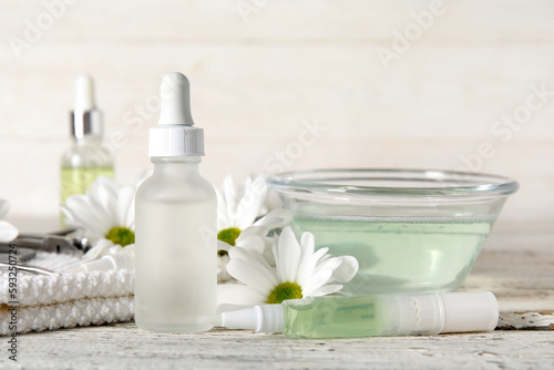 Cuticle oil, bowl of water, towel and chamomile flowers on light wooden background