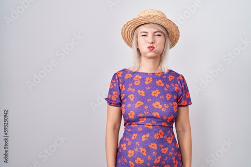 Young caucasian woman wearing flowers dress and summer hat puffing cheeks with funny face. mouth inflated with air, crazy expression.