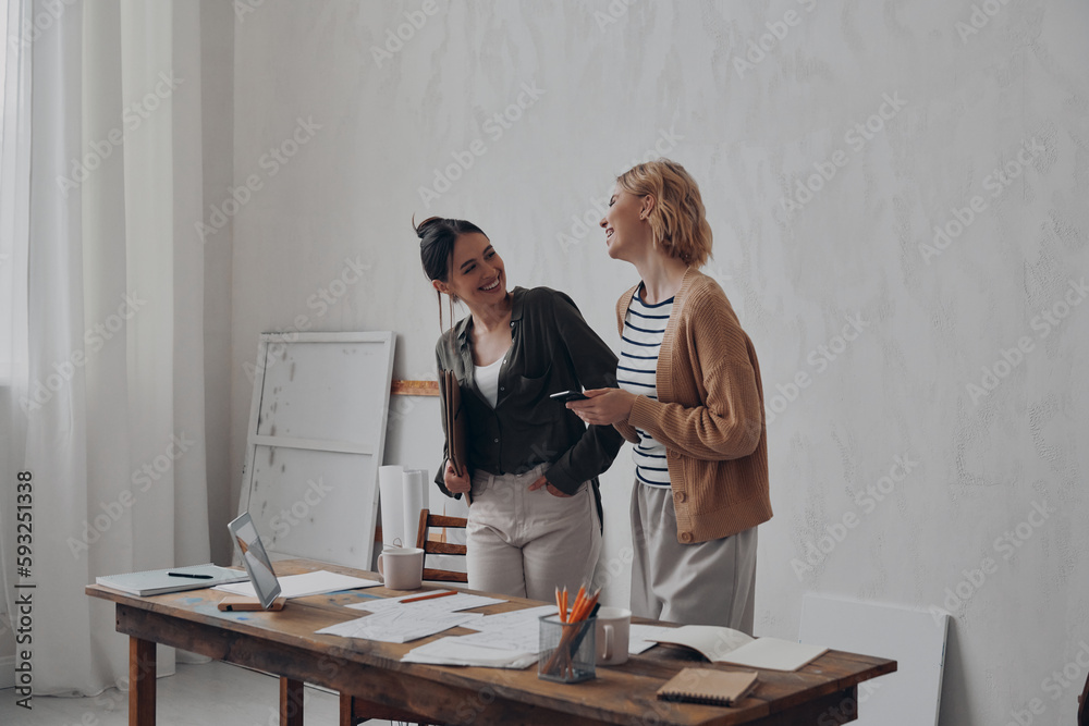 Two cheerful young women talking and smiling while standing near their creative working place Two cheerful young women talking and smiling while standing near their creative working place