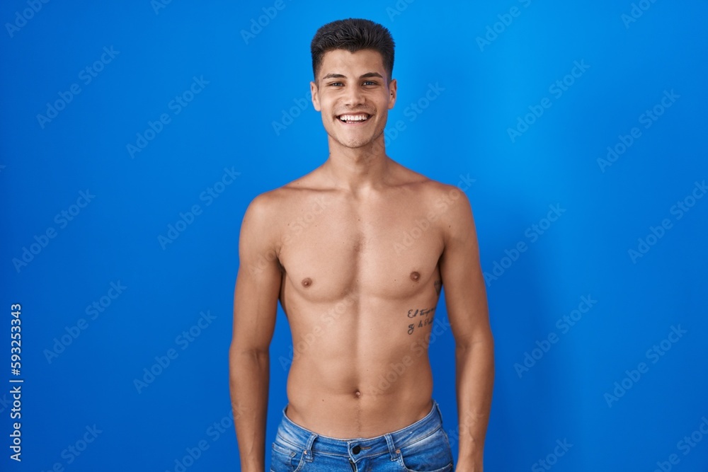 Young hispanic man standing shirtless over blue background with a happy and cool smile on face. lucky person.