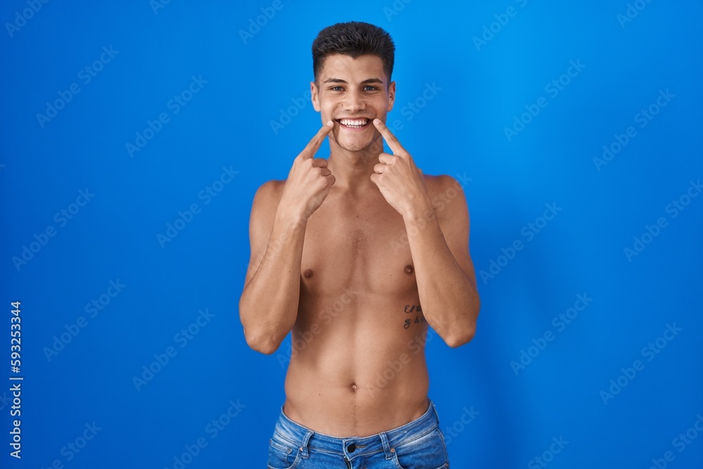 Young hispanic man standing shirtless over blue background smiling with open mouth, fingers pointing and forcing cheerful smile