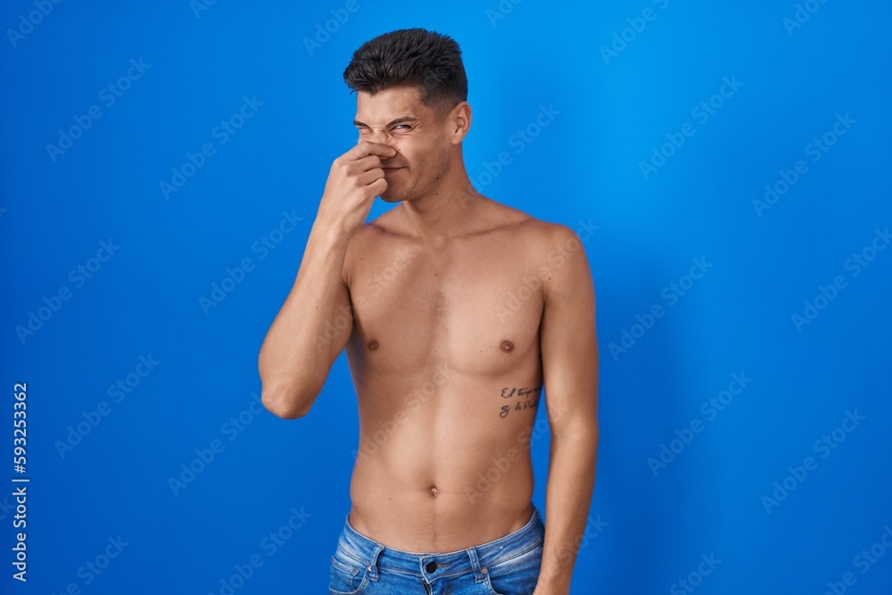 Fototapeta premium Young hispanic man standing shirtless over blue background smelling something stinky and disgusting, intolerable smell, holding breath with fingers on nose. bad smell