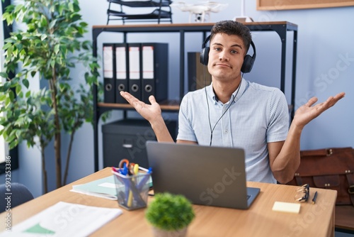 Cuadro en lienzo Young hispanic man working at the office wearing headphones clueless and confused expression with arms and hands raised