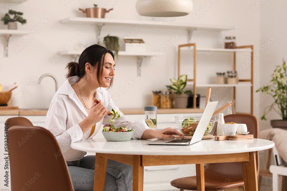 Young woman with laptop eating vegetable salad at table in kitchen