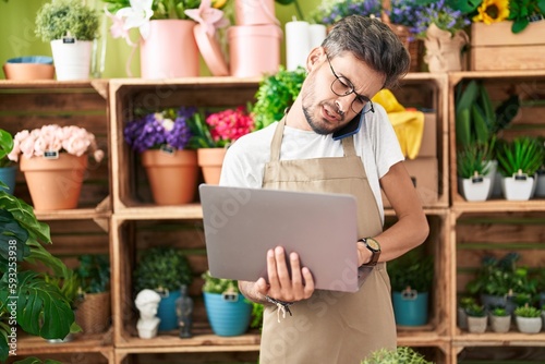 Young hispanic man florist talking on smartphone using laptop at flower shop
