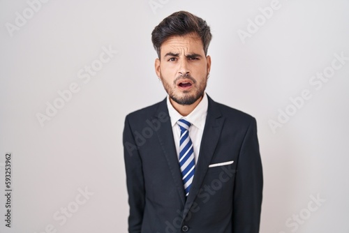 Young hispanic man with tattoos wearing business suit and tie in shock face, looking skeptical and sarcastic, surprised with open mouth
