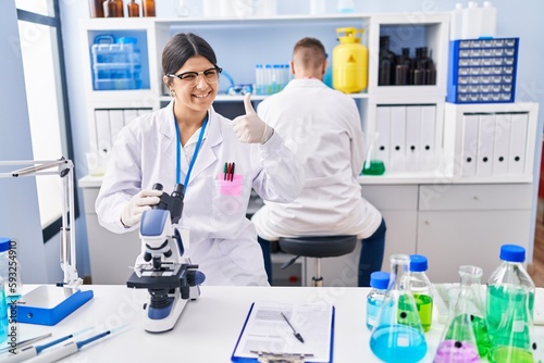 Young woman working at scientist laboratory smiling happy and positive, thumb up doing excellent and approval sign
