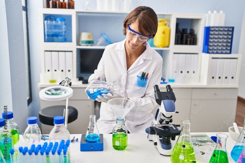 Fototapeta premium Young beautiful hispanic woman scientist pouring liquid on bottle at laboratory