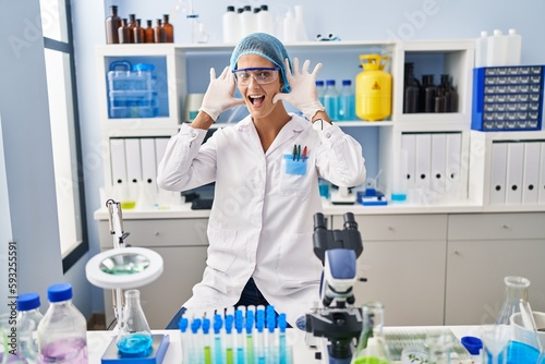 Brunette woman working at scientist laboratory smiling cheerful playing peek a boo with hands showing face. surprised and exited