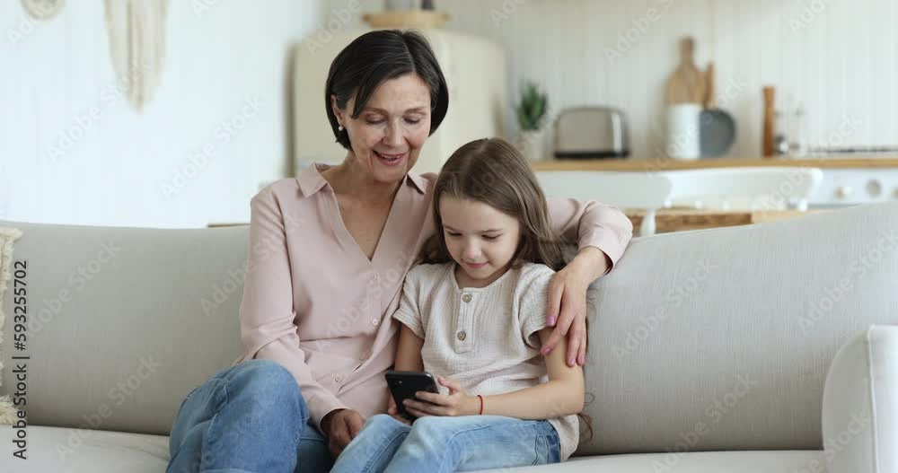 Cheerful pretty middle aged grandma hugging granddaughter girl on sofa. Kid and grandmother using cellphone at home together, playing online game, chatting, looking at display, smiling, laughing Cheerful pretty middle aged grandma hugging granddaughter girl on sofa. Kid and grandmother using cellphone at home together, playing online game, chatting, looking at display, smiling, laughing