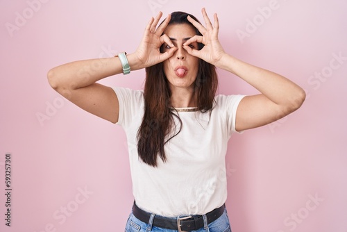 Young brunette woman standing over pink background doing ok gesture like binoculars sticking tongue out, eyes looking through fingers. crazy expression.