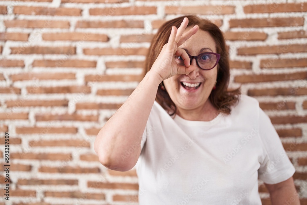 Senior woman with glasses standing over bricks wall doing ok gesture with hand smiling, eye looking through fingers with happy face.