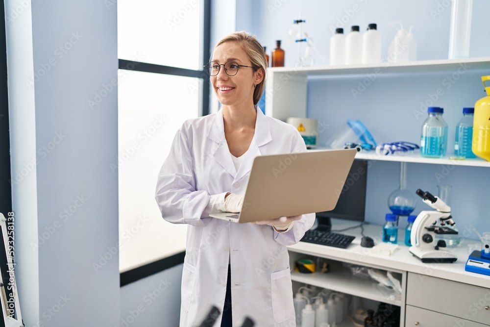 Young blonde woman scientist smiling confident using laptop at laboratory