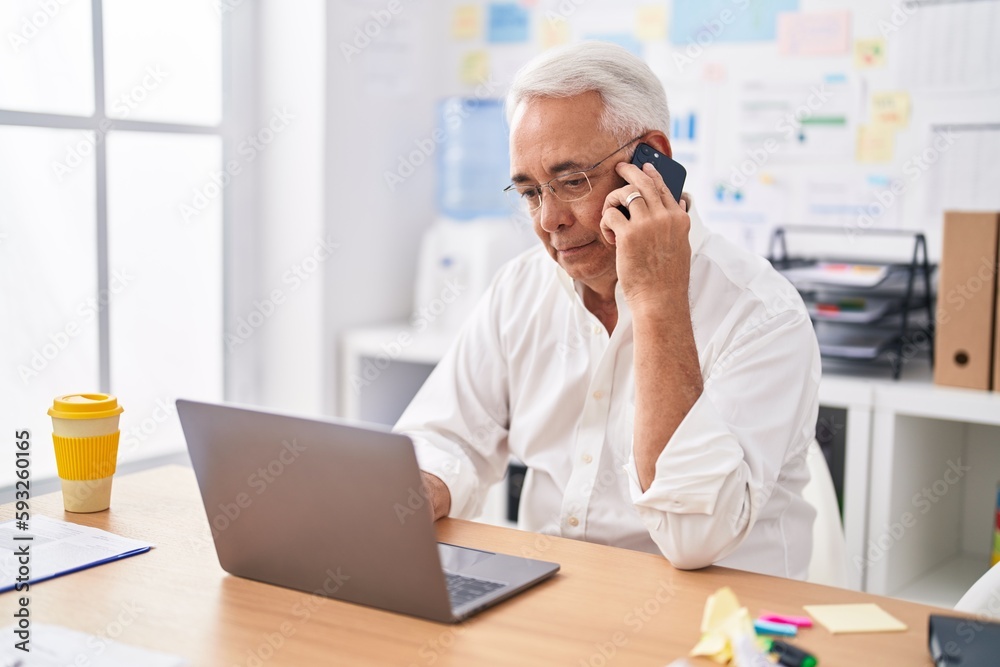 Middle age grey-haired man business worker using laptop talking on smartphone at office Middle age grey-haired man business worker using laptop talking on smartphone at office