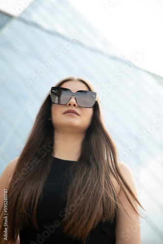 Closeup image of a graceful girl with long hair and makeup in black top standing behind blurred building and bright background.