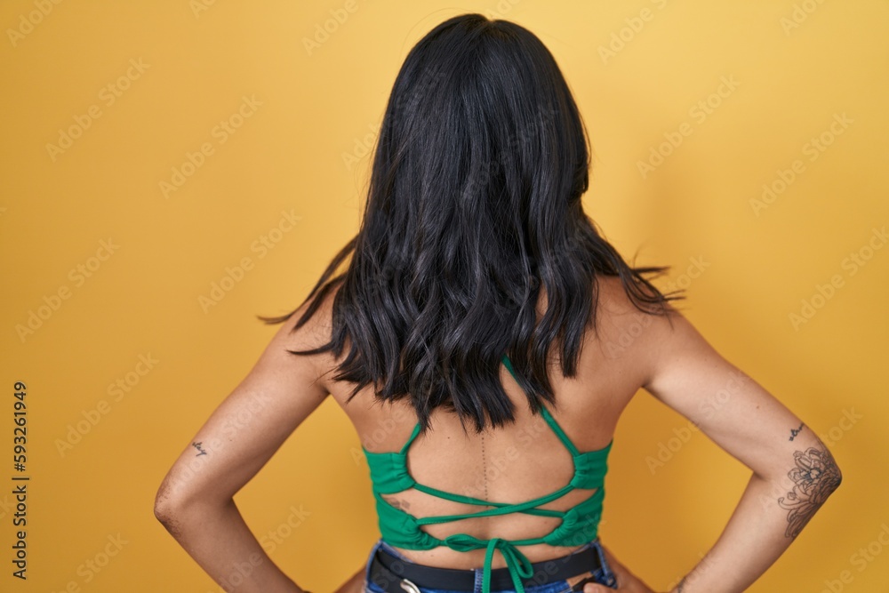 Brunette woman standing over yellow background standing backwards looking away with arms on body Brunette woman standing over yellow background standing backwards looking away with arms on body