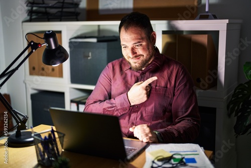 Plus size hispanic man with beard working at the office at night cheerful with a smile of face pointing with hand and finger up to the side with happy and natural expression on face