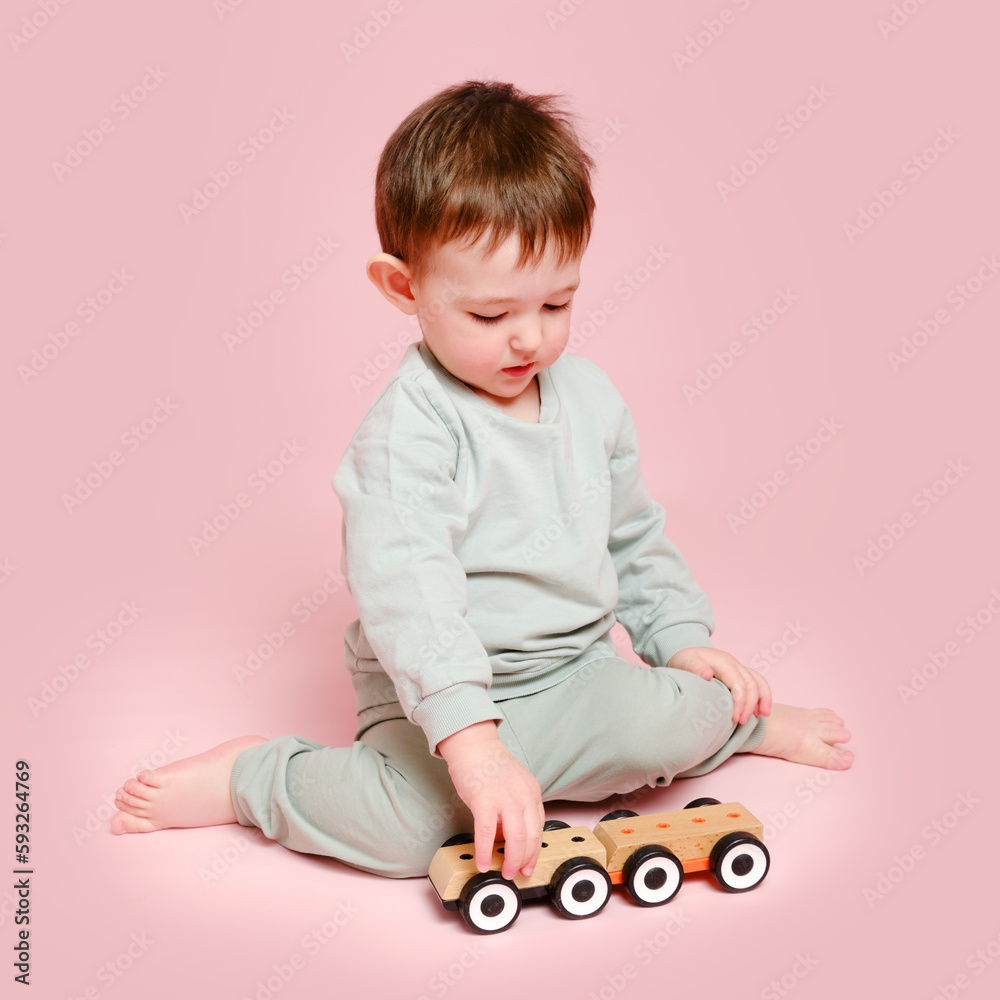 Naklejka premium Happy toddler baby is playing with a toy car against a pink background. Child boy rolls a wooden toy car. Kid age one year eight months, full height