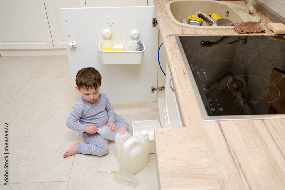 A child is playing with chemical cleaning products under the sink in the kitchen. Baby holds bottles with detergent. Kid aged about two years (one year nine months) A child is playing with chemical cleaning products under the sink in the kitchen. Baby holds bottles with detergent. Kid aged about two years (one year nine months)